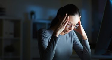 Young businesswoman with headache touching her head while sitting in front of computer monitor by her workplace at night