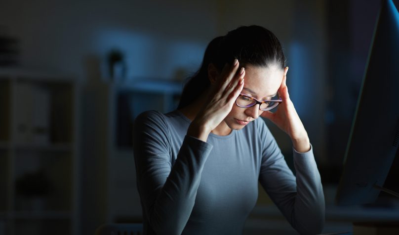 Young businesswoman with headache touching her head while sitting in front of computer monitor by her workplace at night