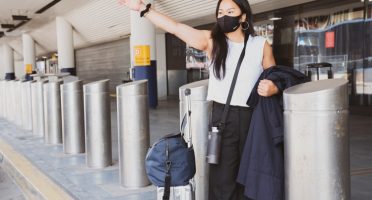 Na imagem, uma mulher com máscara e malas acena, pedindo um táxi, em frente a um aeroporto.