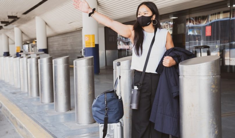 Na imagem, uma mulher com máscara e malas acena, pedindo um táxi, em frente a um aeroporto.